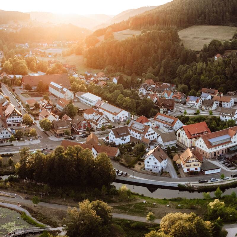 Eine malerische Kleinstadt in einer hügeligen Landschaft mit dicht aneinander gereihten Häusern und einem kleinen Teich im Vordergrund wird bei Sonnenuntergang beleuchtet.