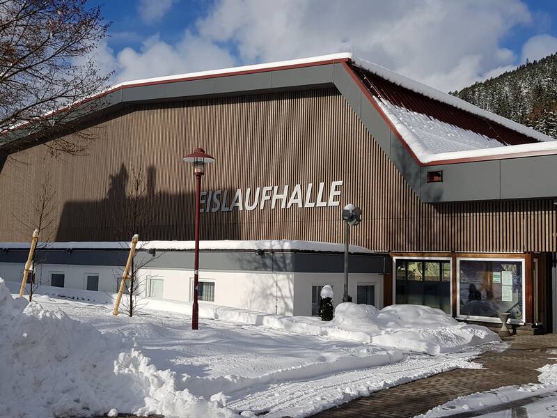 Eine mit Schnee bedeckte Eislaufhalle steht unter einem teils bewölkten Himmel, umgeben von winterlicher Landschaft.