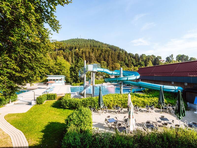 Ein Schwimmbad im Freien mit einer blauen Wasserrutsche, umgeben von grüner Vegetation und Liegestühlen, mit einem bewaldeten Hügel im Hintergrund.
