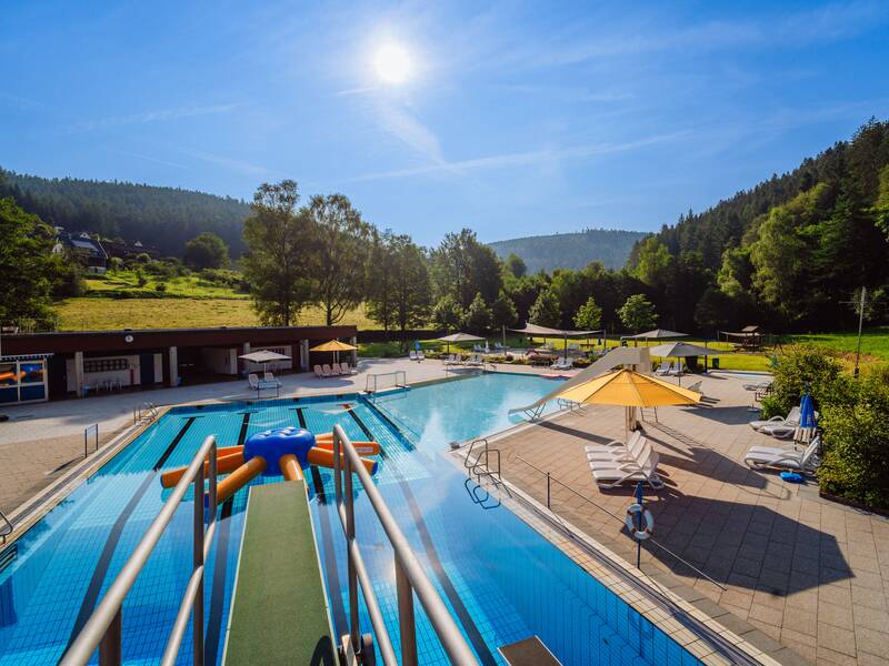 Ein Freibad bei sonnigem Wetter mit Liegestühlen, Sonnenschirmen und umliegenden bewaldeten Hügeln.
