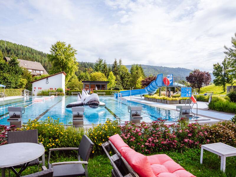 Ein Freibad mit einem großen aufblasbaren Hai im Wasser, umgeben von Bäumen und einer Rutsche im Hintergrund, mit Sitzgelegenheiten und Blumen im Vordergrund.