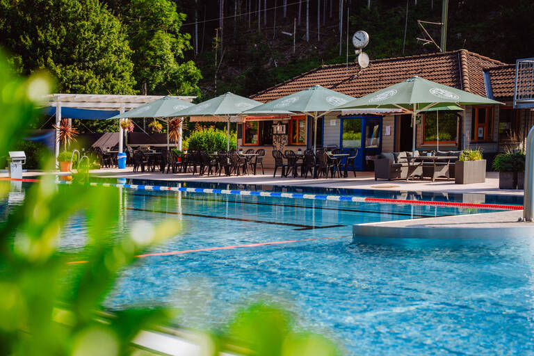 Ein Freibad mit einem klaren Schwimmbecken, umgeben von Sonnenschirmen, Tischen und Stühlen vor einem Gebäude mit Ziegeldach und üppiger Vegetation im Hintergrund.