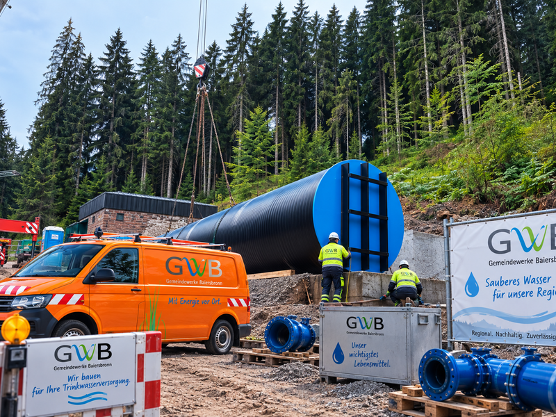 Ein großer schwarzer Tank wird mit einem Kran in einer waldreichen Umgebung installiert, während Arbeiter in Sicherheitskleidung von der Gemeindewerke Baiersbronn anwesend sind.